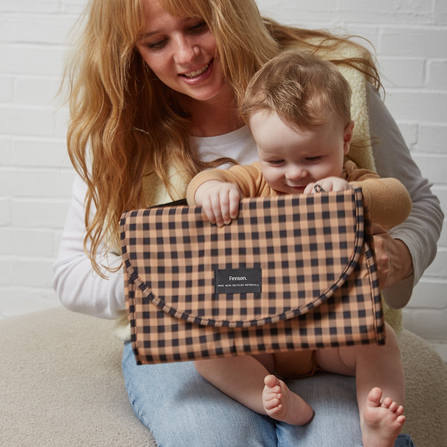 A woman sitting on a beige floor cushion holds a baby while presenting the Oslo gingham check patterned travel changing mat. The woman is wearing a cream vest over a white long-sleeve shirt and blue jeans.