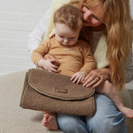 A woman and a baby sit together on a beige sofa against a white brick wall, with the woman holding a mocha textured borg changing mat across her lap.