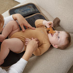 A baby in a tan long-sleeve bodysuit lies on a beige sofa while an adult adjusts the baby's diaper. A changing mat with mesh pockets is visible behind them.