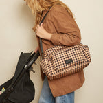 A woman in a brown jacket and blue jeans carries a gingham checked diaper bag over her shoulder while pushing a black stroller against a plain beige wall.