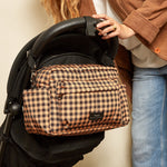 A person in a brown jacket and blue jeans holds a black stroller with a brown and beige gingham diaper bag attached to the side, against a plain beige wall.