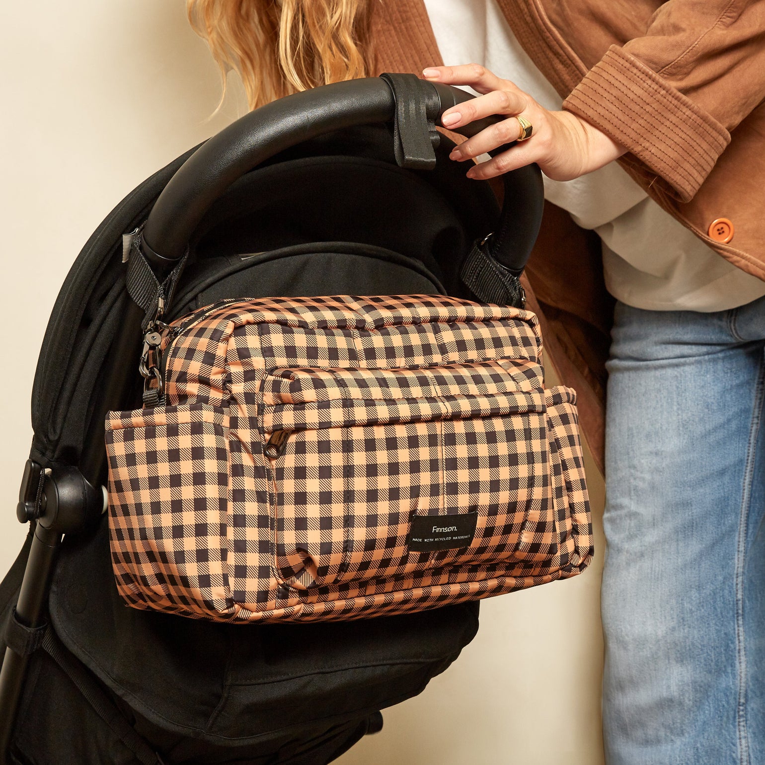 A woman in a brown jacket and blue jeans holds a black stroller with Freya gingham check patterned stroller organiser bag attached to the side, against a plain beige wall.