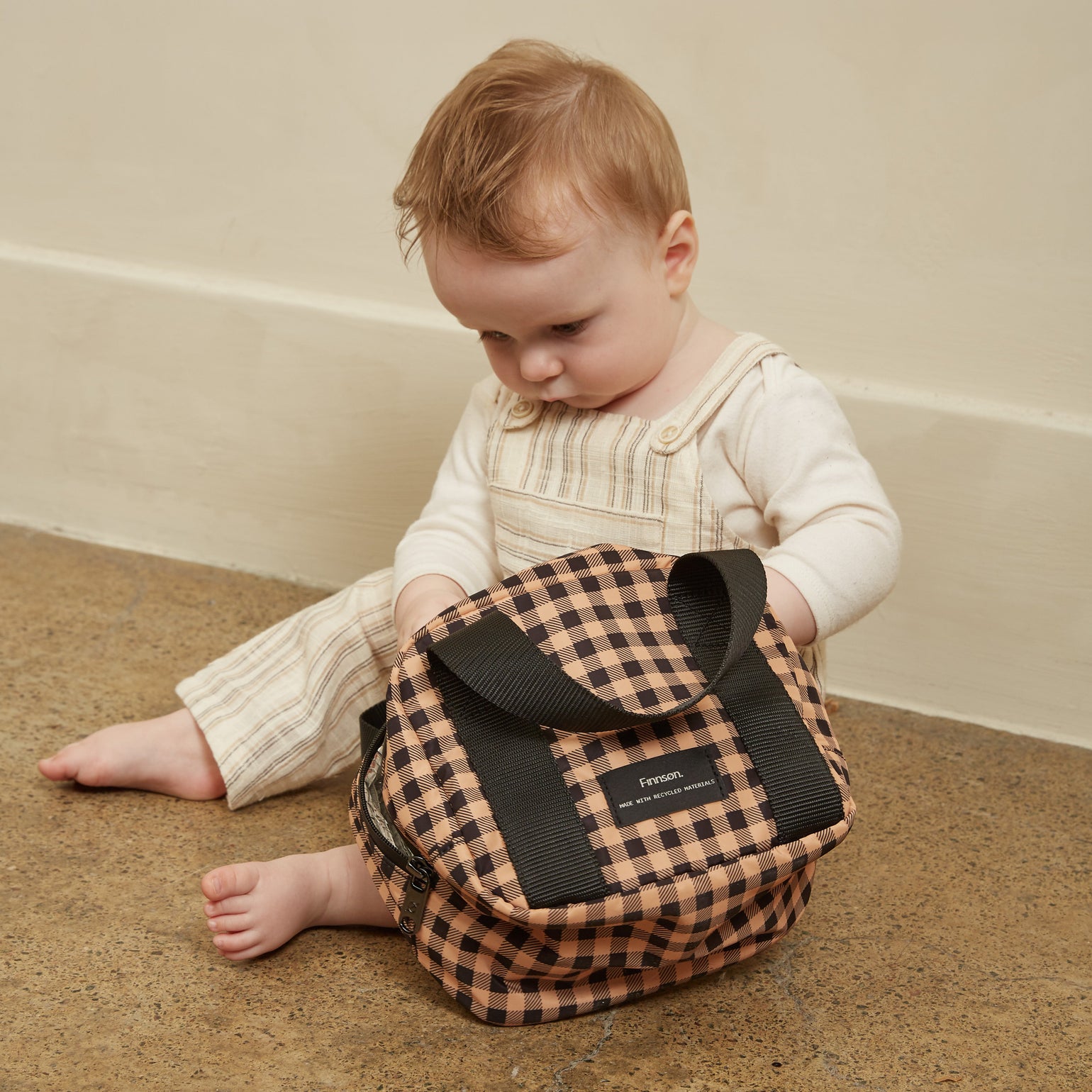 A baby sitting on a textured floor next to the Ebba gingham check patterned lunch/snack cool bag. The baby is wearing cream-colored overalls and a long-sleeve top.
