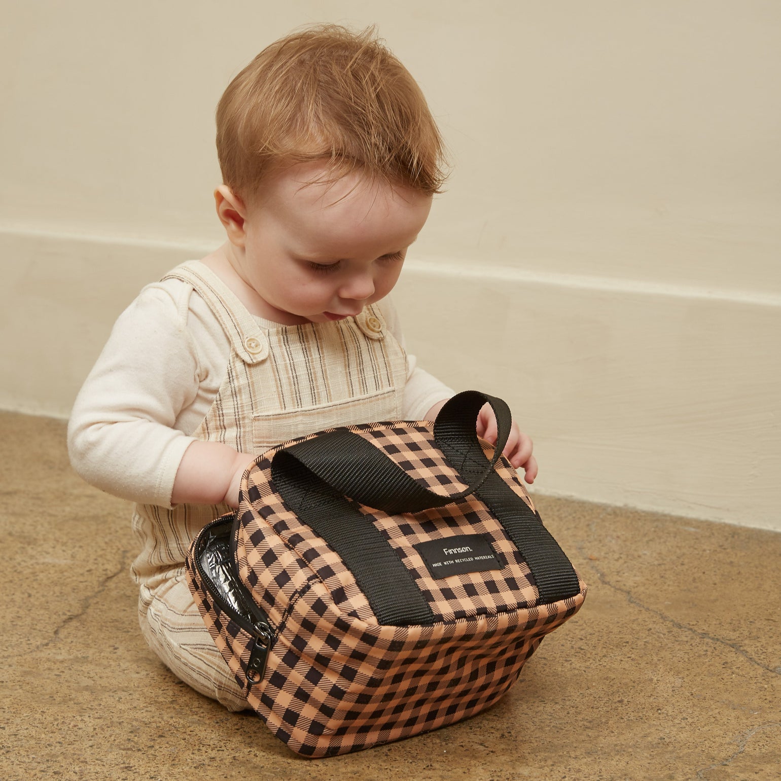 A baby sitting on a concrete floor against a beige wall, holding the Ebba gingham check patterned lunch/snack cool bag.
