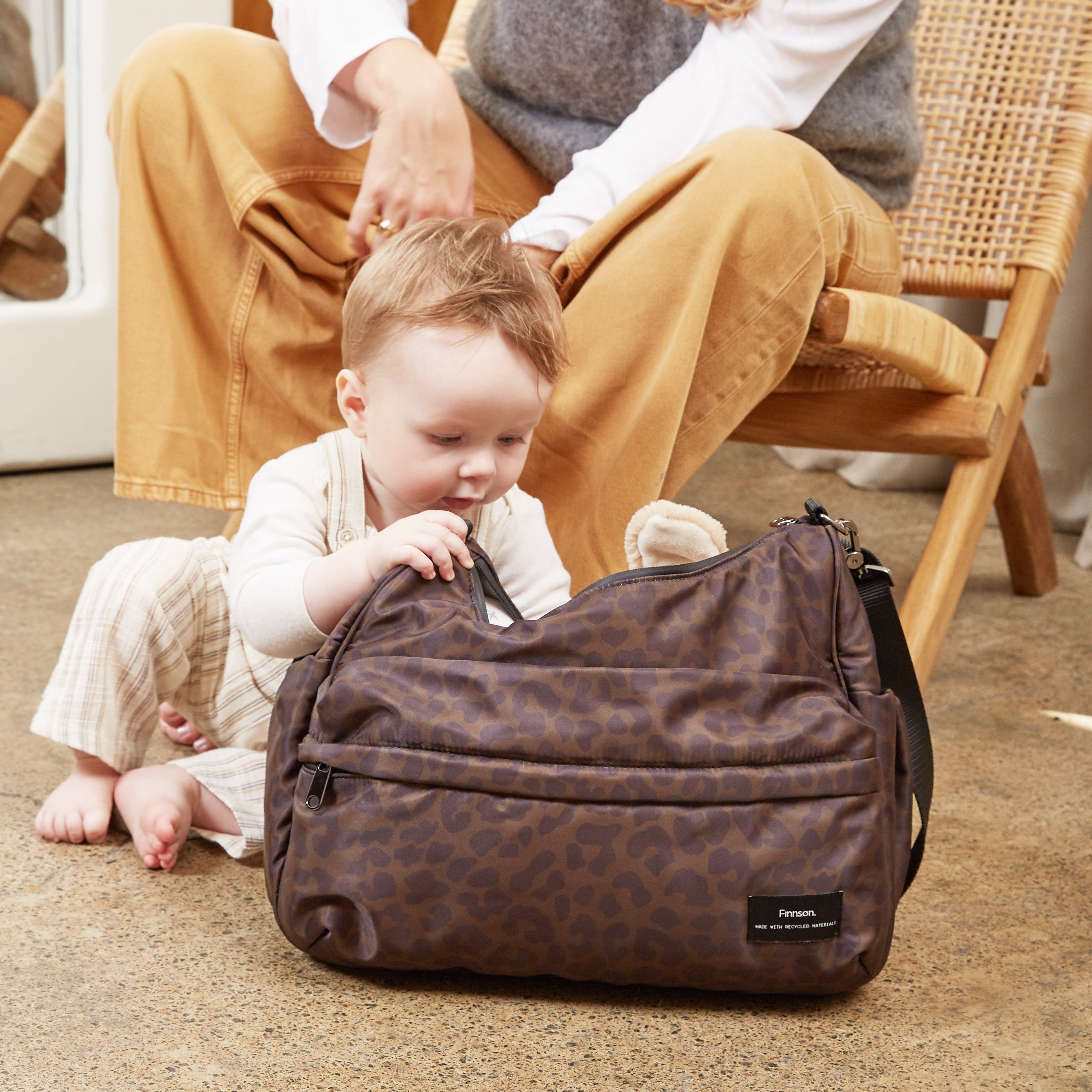 A woman and a baby sit on a carpeted floor in a warm-toned room, with the woman adjusting the baby's outfit and the baby touching the Clara chocolate leopard diaper bag in the foreground.