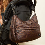 A woman in a tan jacket and jeans leans over a black stroller, the clara chocolate brown changing bag is hanging from the stroller handle.