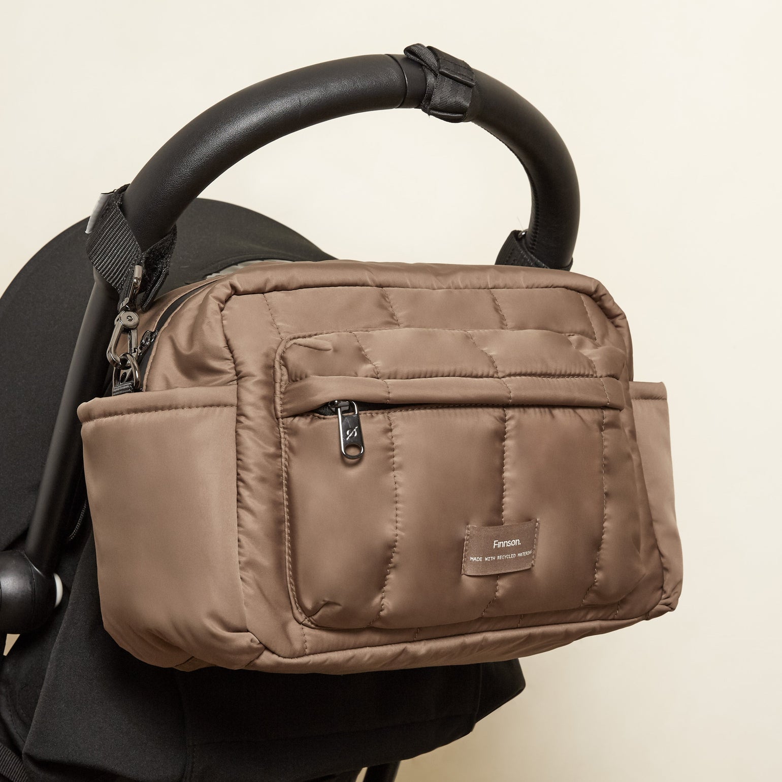 A mocha nylon diaper bag with a front zippered pocket and a small brand label, shown hanging from a black stroller handle against a plain beige wall.