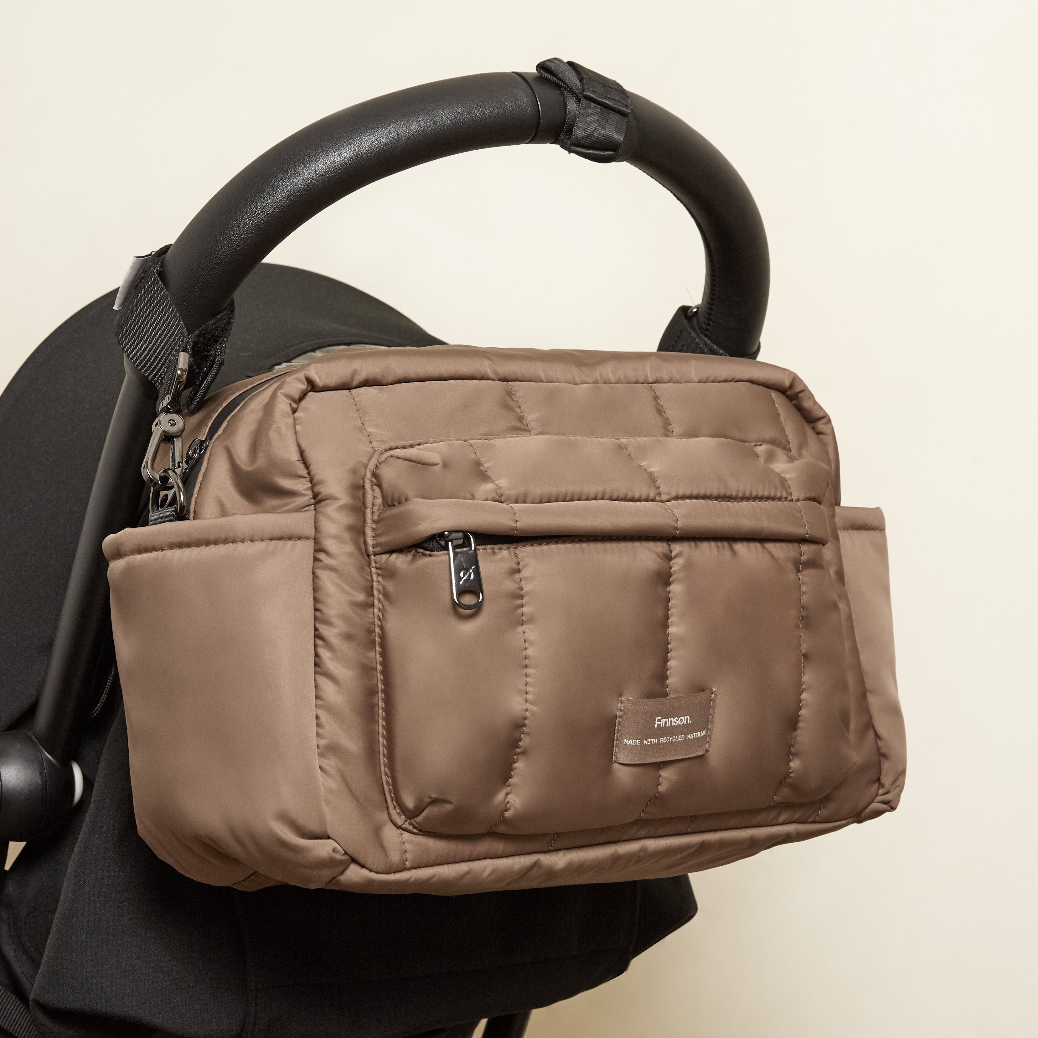 A mocha nylon diaper bag with a front zippered pocket and a small brand label, shown hanging from a black stroller handle against a plain beige wall.