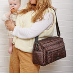 A woman with long blonde hair holds a baby while wearing a leopard print  patterned crossbody diaper bag. She is wearing a cream-colored fuzzy vest over a white long-sleeve shirt and tan pants, standing against a white brick wall.