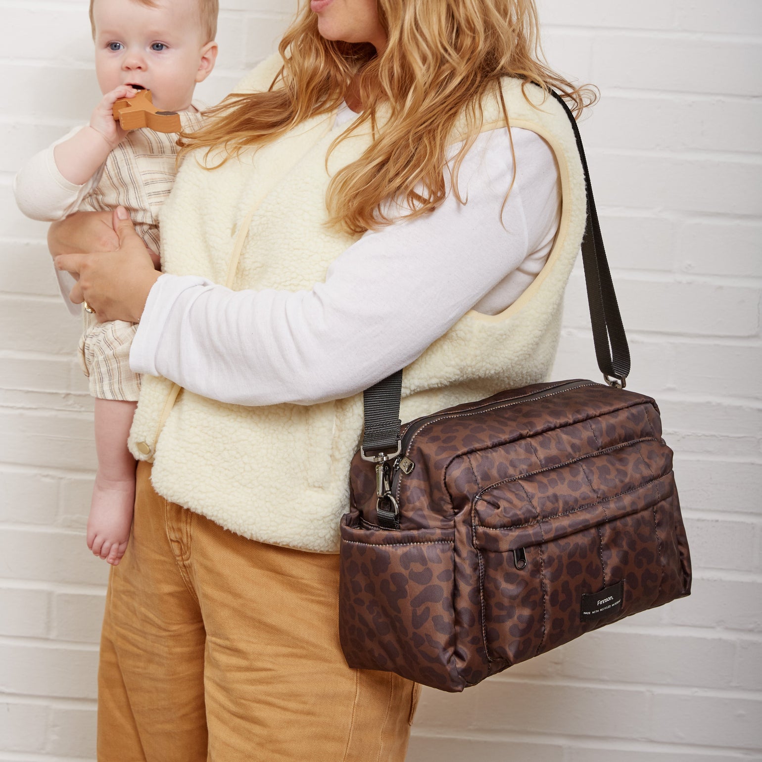 A woman with long blonde hair holds a baby while wearing the Freya choco leopard print stroller organizer bag. She is wearing a cream-colored fuzzy vest over a white long-sleeve shirt and tan pants, standing against a white brick wall.