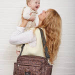 A woman holding a baby wearing a leopard-print crossbody bag across her body, set against a white brick wall.