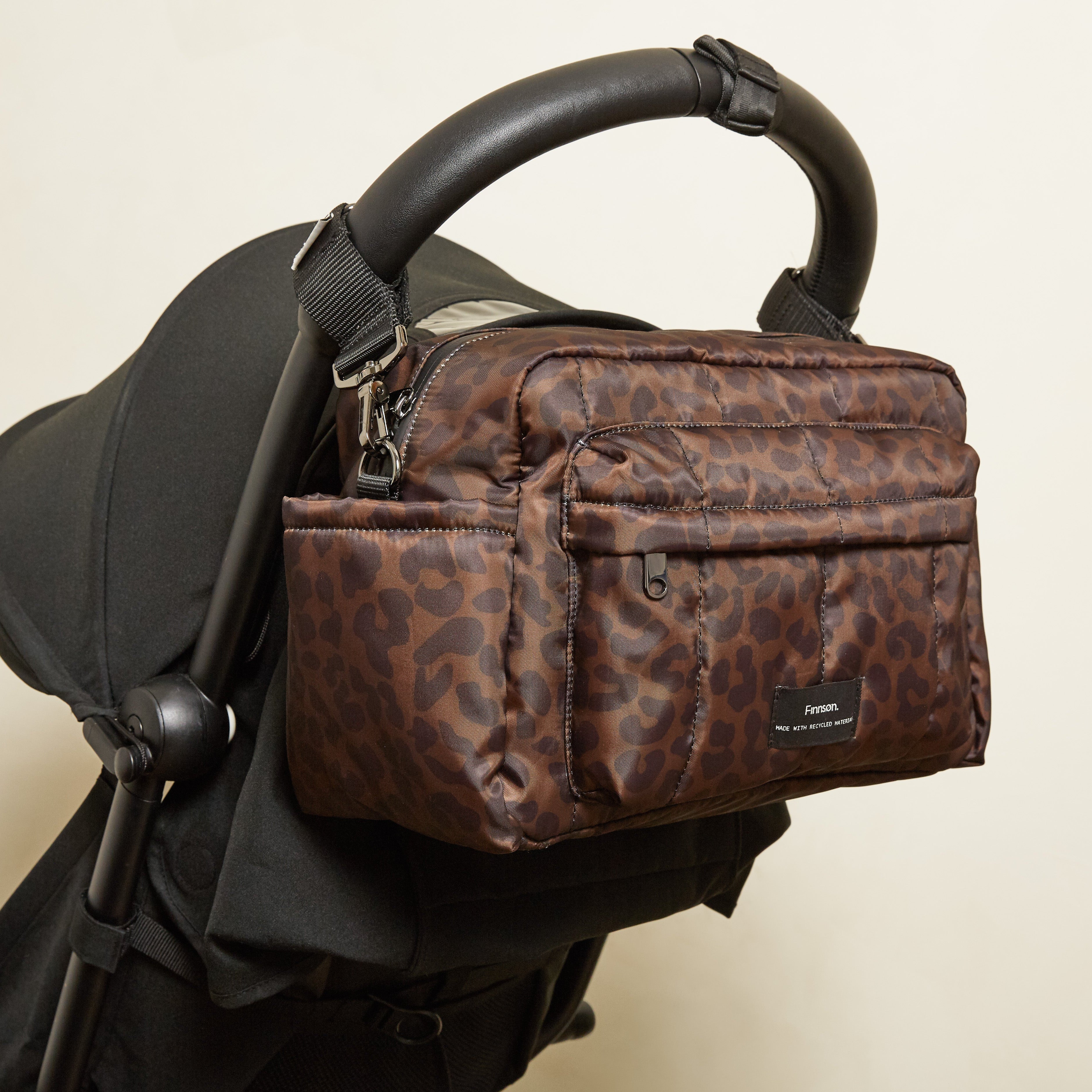 A brown leopard-print stroller bag hanging from a black stroller handle against a plain light beige wall.
