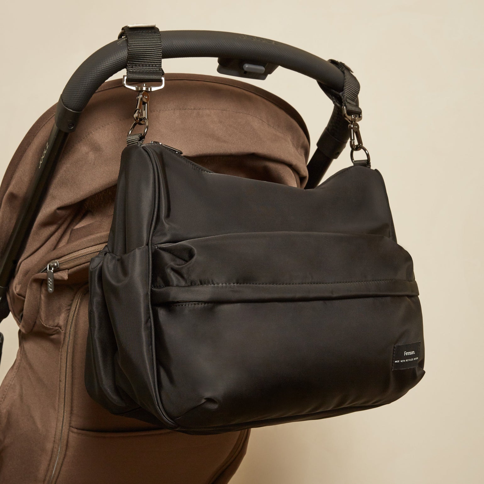 The Finnson Clara changing bag with a front zippered pocket and side gusset, shown hanging from the handle of a brown stroller against a plain beige wall.