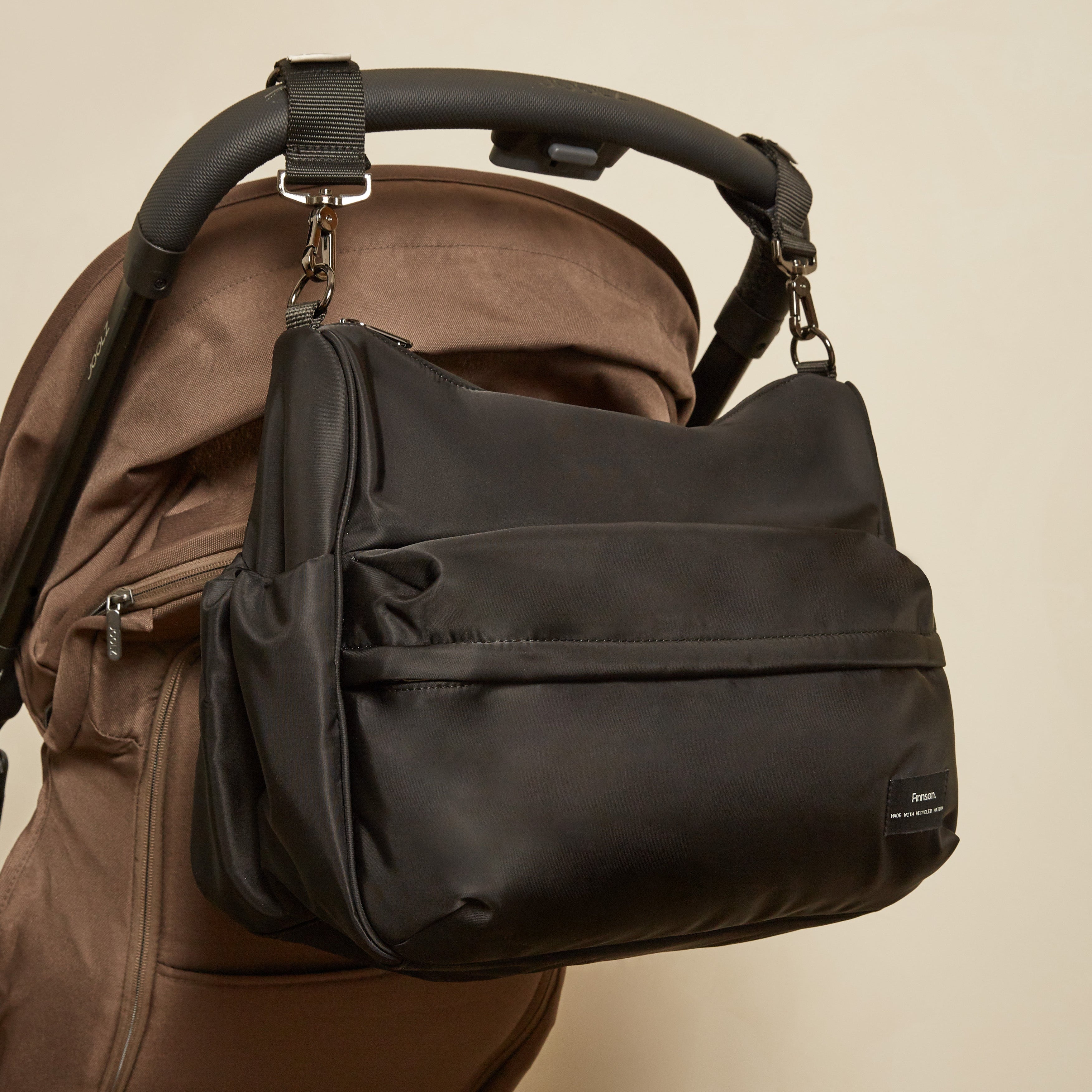 The Finnson Clara changing bag with a front zippered pocket and side gusset, shown hanging from the handle of a brown stroller against a plain beige wall.