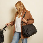 A woman with blonde hair stands against a beige wall, wearing a brown coat, white t-shirt, and blue jeans. She is holding the Clara Finnson changing bag with one hand and carrying a large black messenger bag on her shoulder with the other.