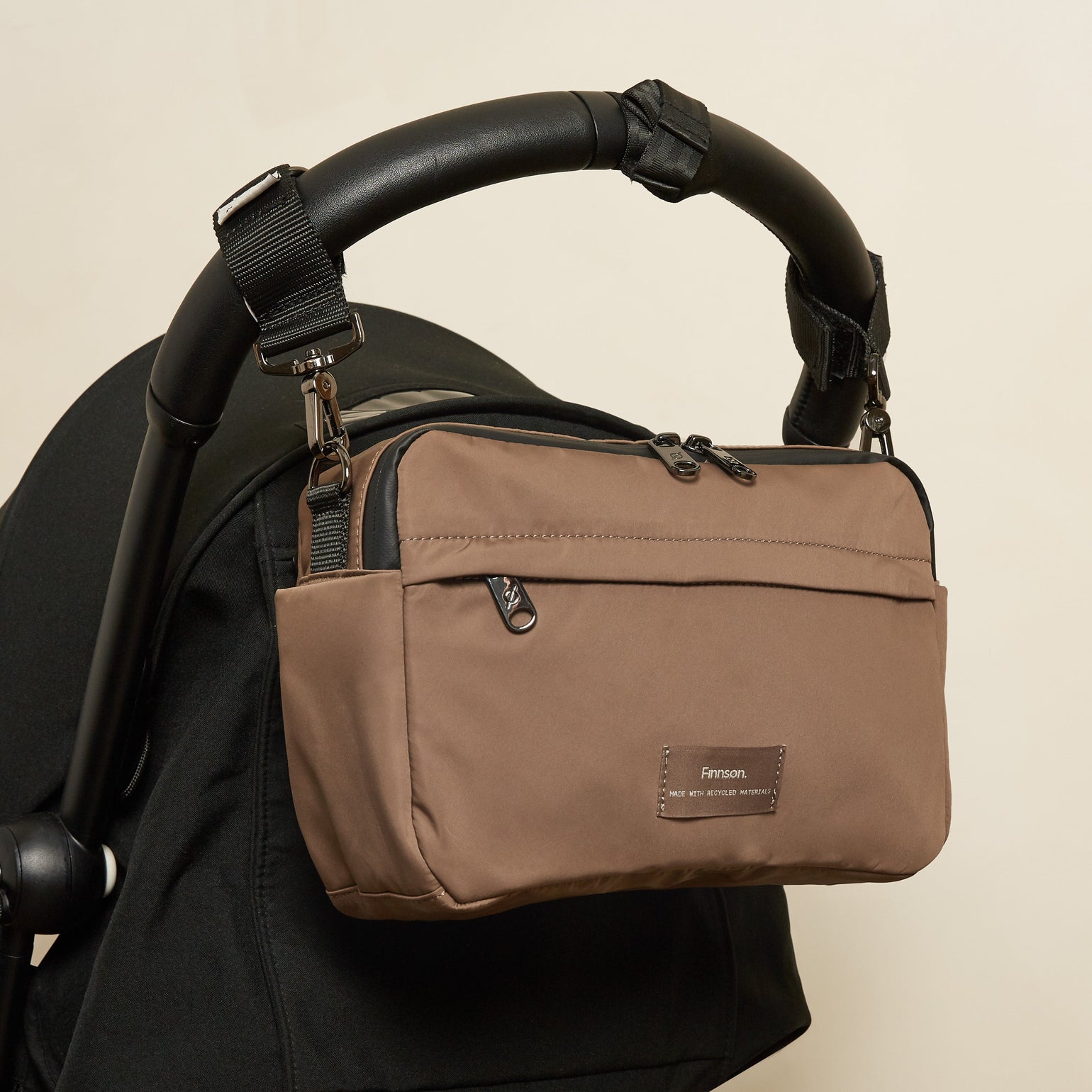 A Frida mocha eco stroller organiser bag, shown hanging from a black stroller handle against a light beige background.