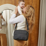 A woman wearing a cream vest and tan pants carrying the Fern eco stroller organiser while holding a baby in front of a wooden wardrobe and mirror.
