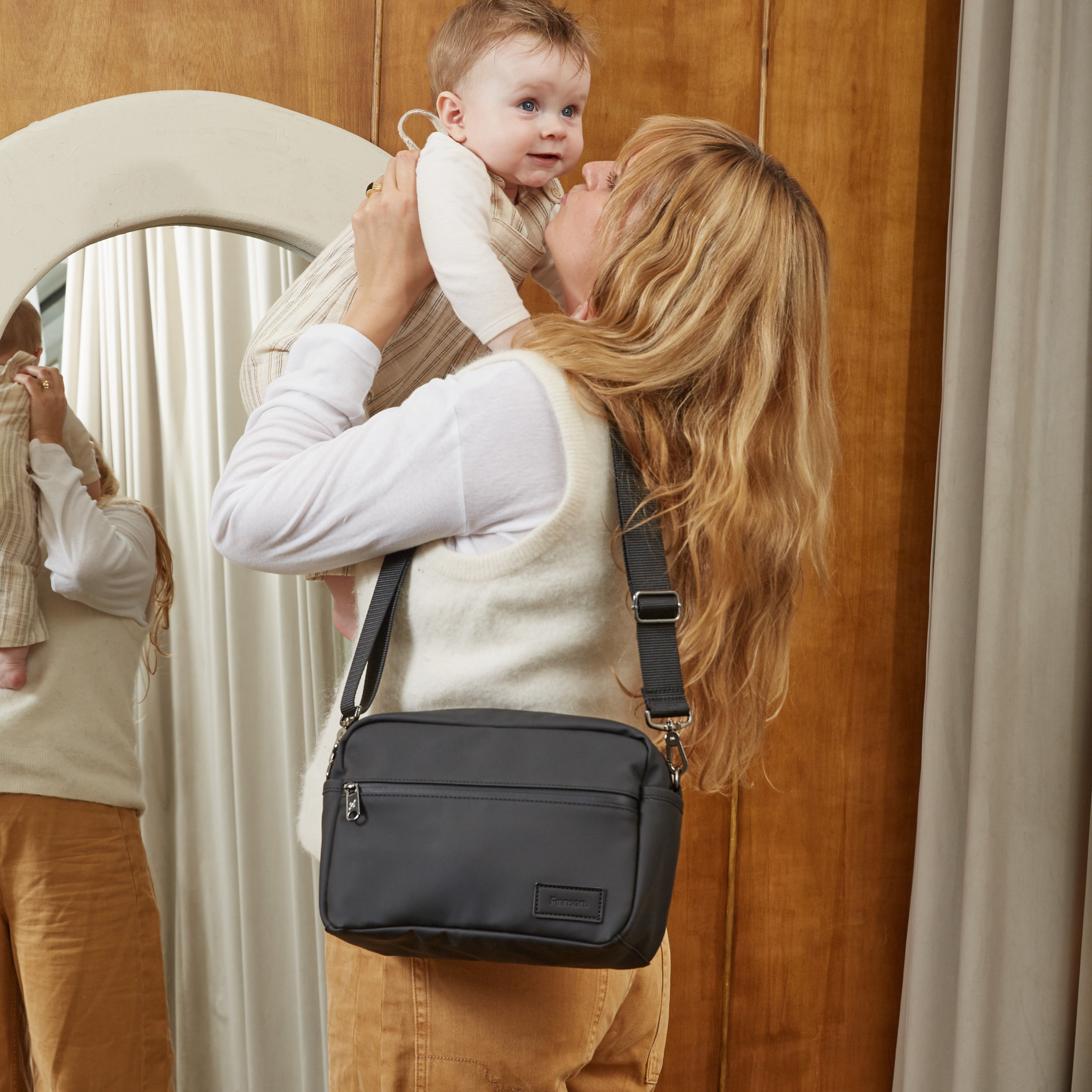 A woman wearing a cream vest and tan pants carrying the Fern eco stroller organiser while holding a baby in front of a wooden wardrobe and mirror.