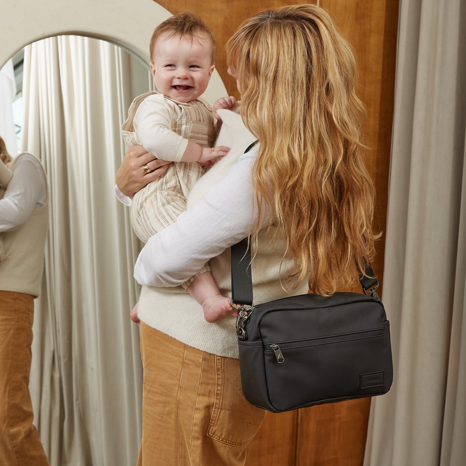 A woman carrying a baby over her shoulder while wearing the Finnson Fern eco stroller organiser. The woman is seen from behind, holding a smiling baby in a cream knit sweater and tan pants, standing in front of a wooden wall and a mirror.