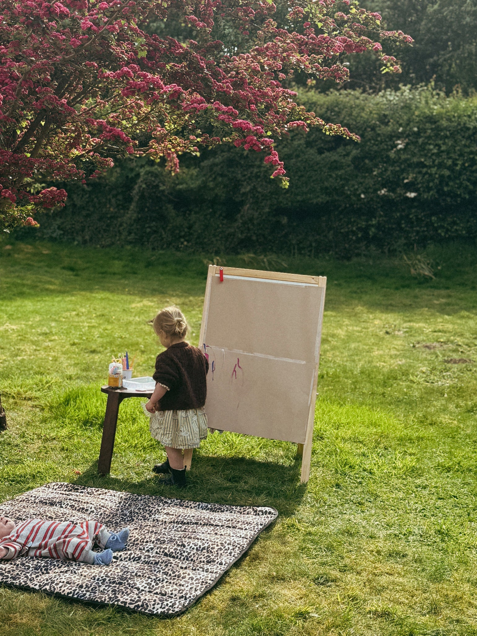 Child painting on an easel outdoors in a garden with the baby resting in the Thoren outdoor/indoor playmat leopard print pattern